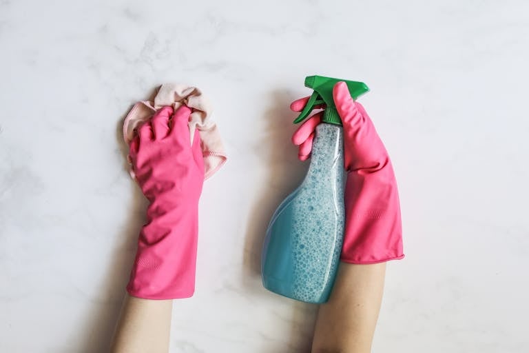 A pair of hands in pink gloves are cleaning with a spray bottle on a white background.